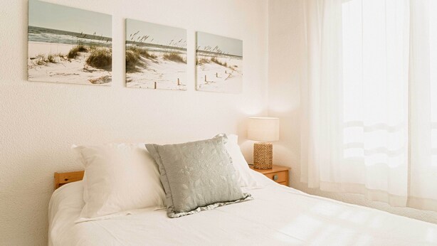 Una cama bien hecha con sábanas blancas y almohada gris, una lámpara de noche y tres obras de arte con motivos playeros.