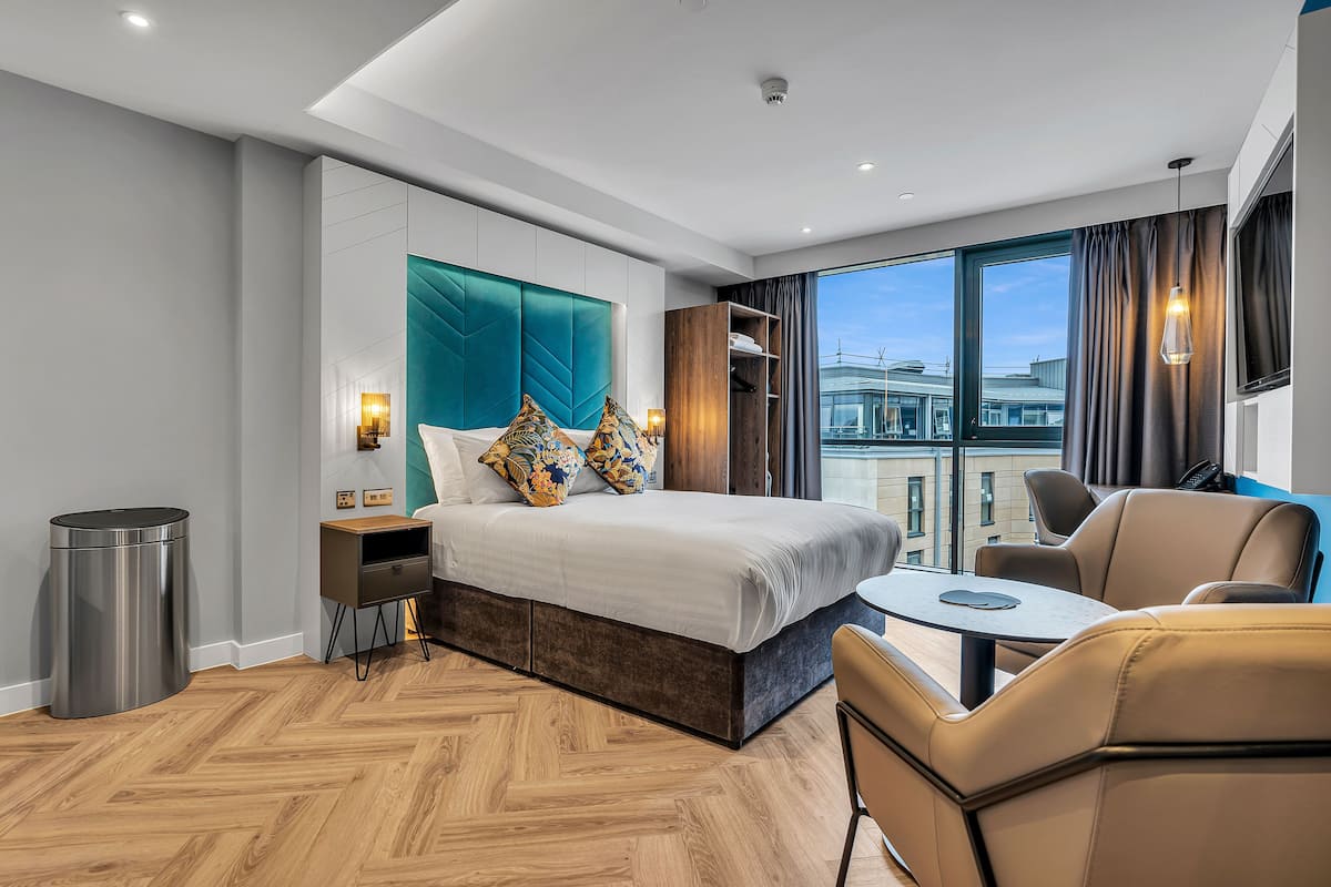 A modern hotel room with a large bed, a small table, a sofa, and a view of the city through large windows.