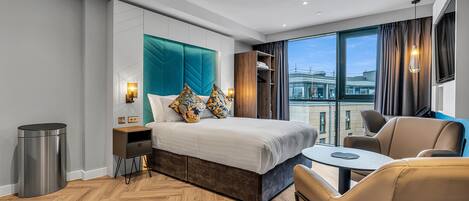 A modern hotel room with a large bed, a small table, a sofa, and a view of the city through large windows.