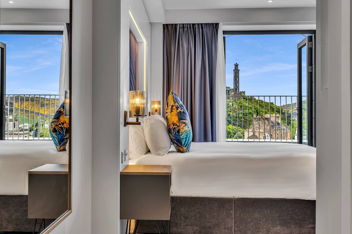 A modern hotel room with a large bed, bedside lamps, and a view of a cityscape and tower through the window.