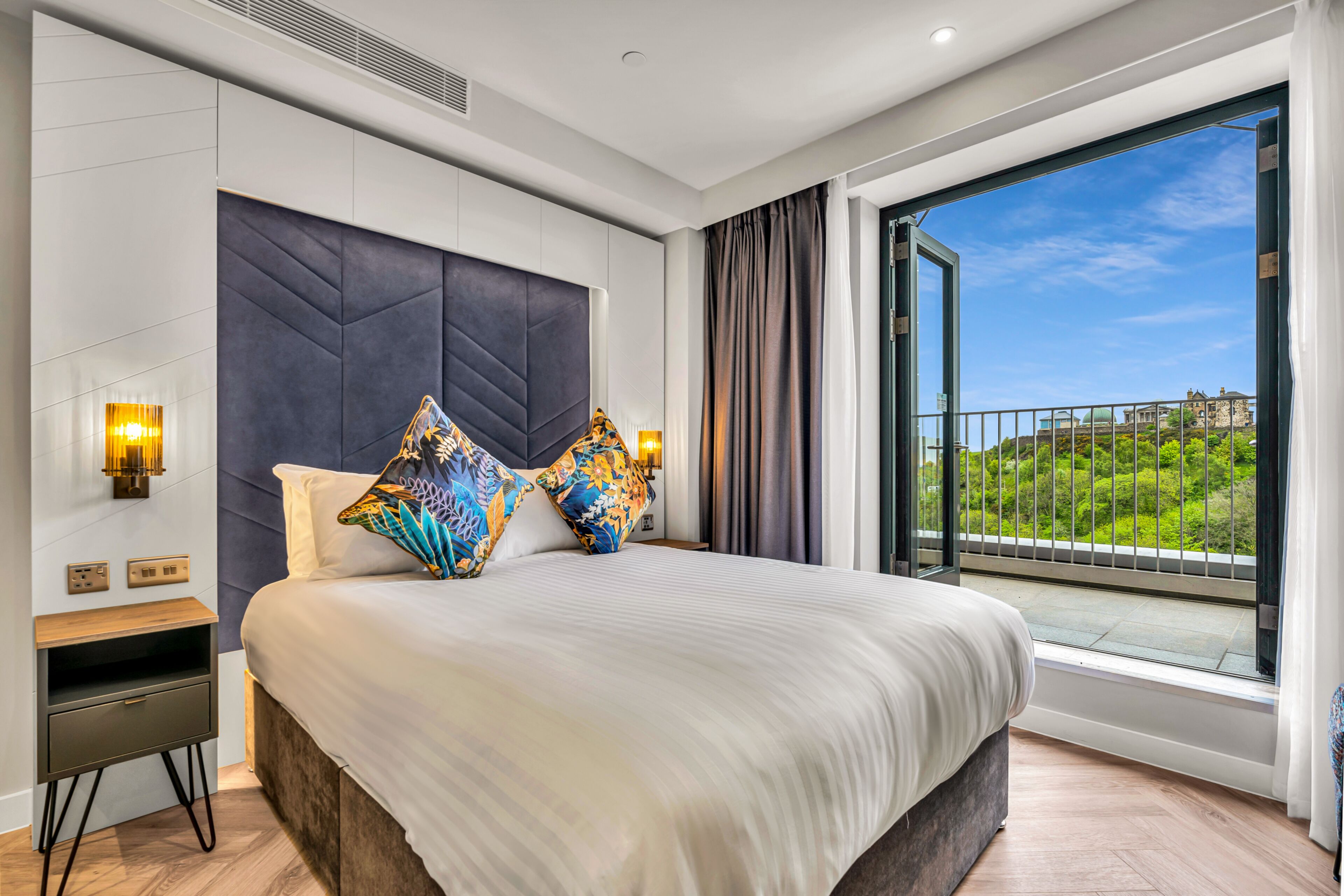 A modern hotel bedroom with a large bed, a nightstand, a wall-mounted TV, and a balcony with a scenic view.