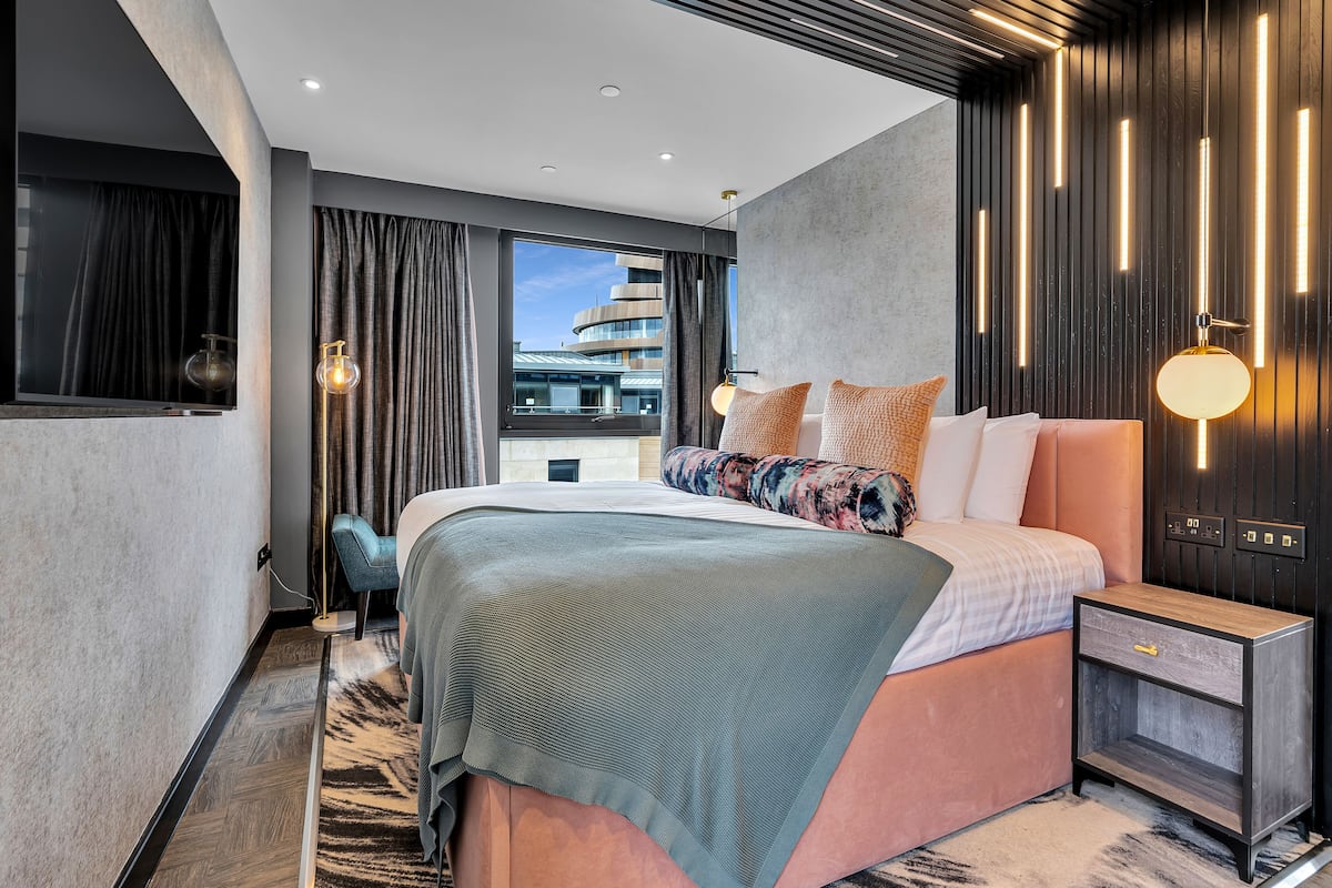 A modern bedroom with a large bed, a nightstand, a desk lamp, and a view of a building through the window.