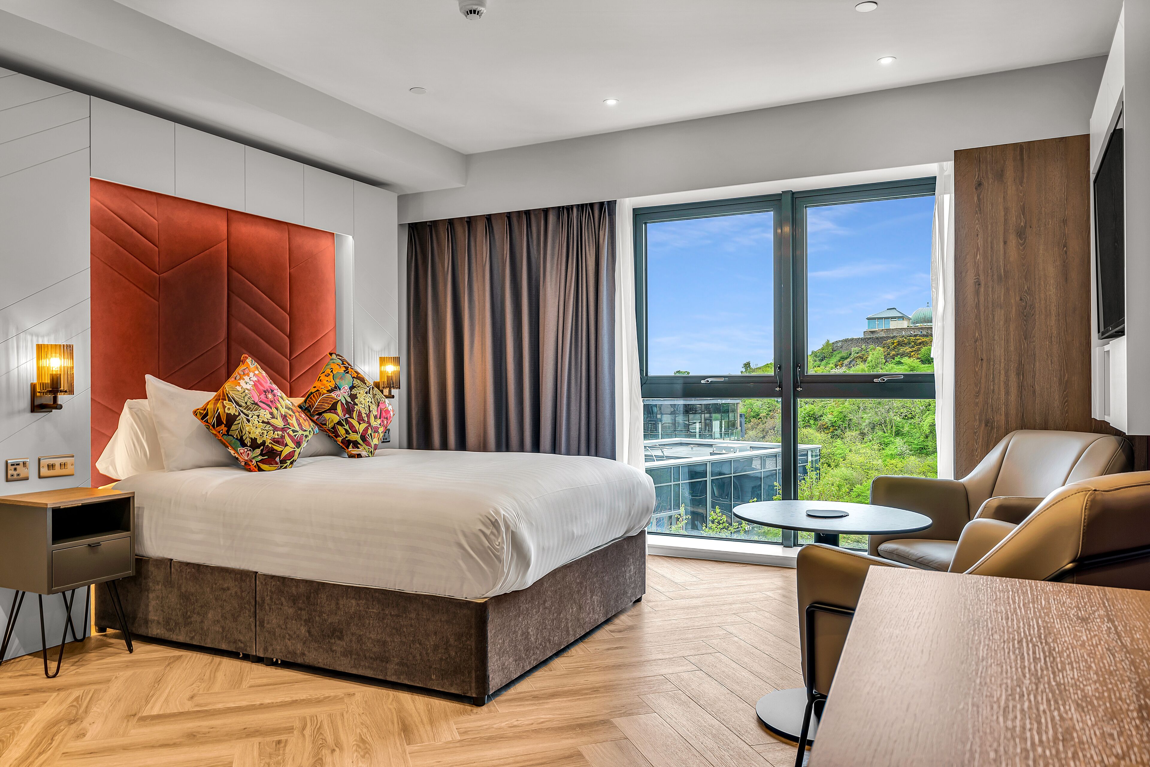 A modern hotel room with a large bed, a desk, a chair, and a view of a cityscape.