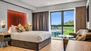 A modern hotel room with a large bed, a desk, a chair, and a view of a cityscape.