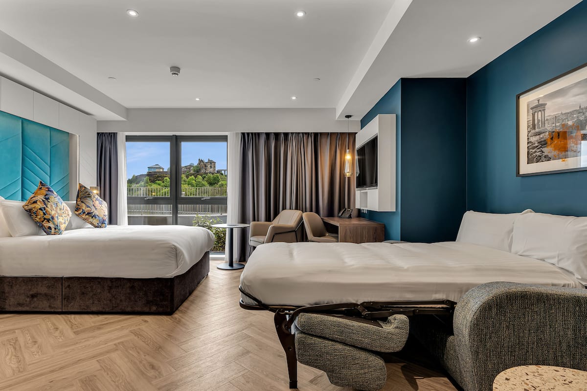 A modern hotel room with two beds, a desk, and a view of a building through large windows.