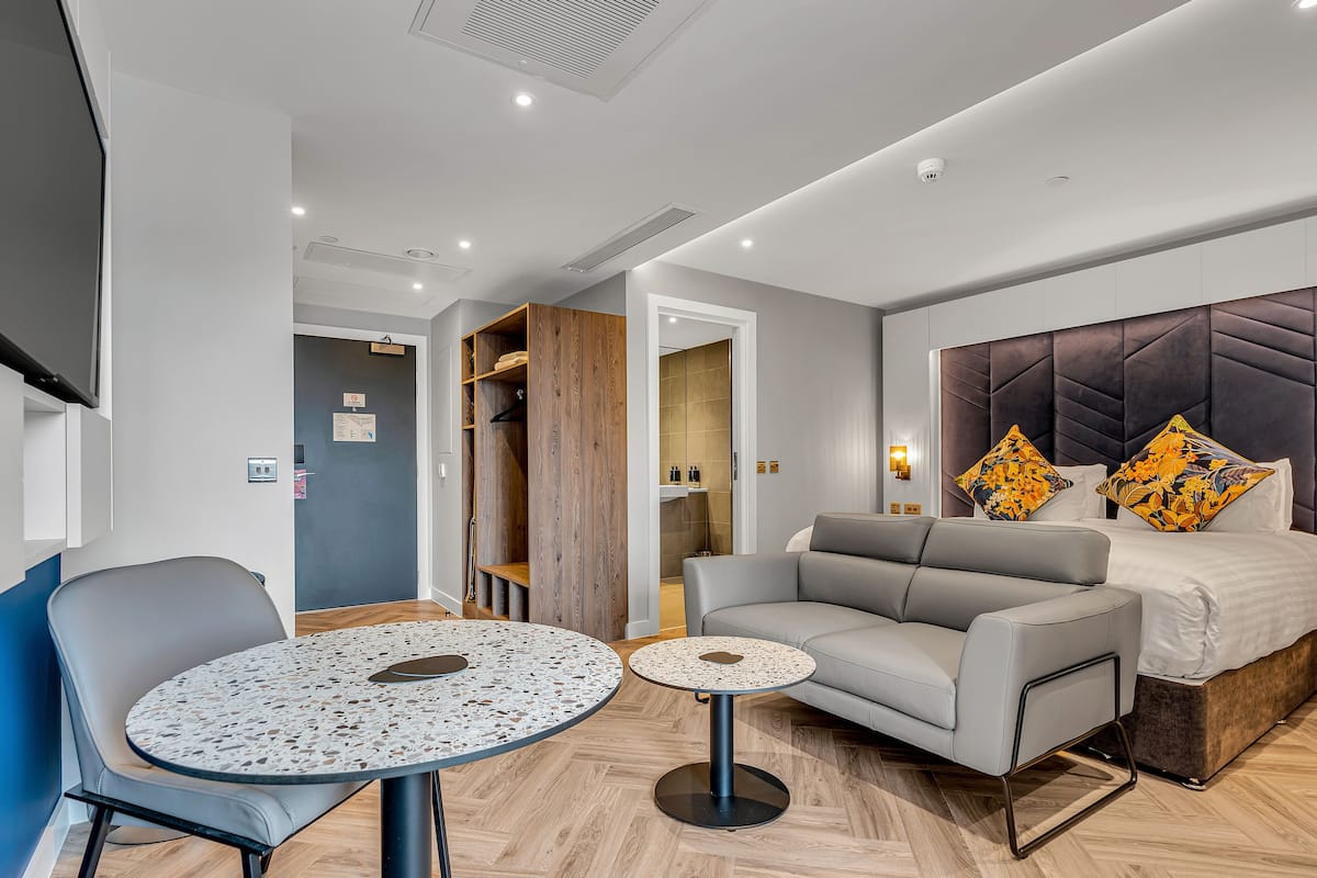 A modern hotel room with a bed, a grey sofa, a round table, and a chair.