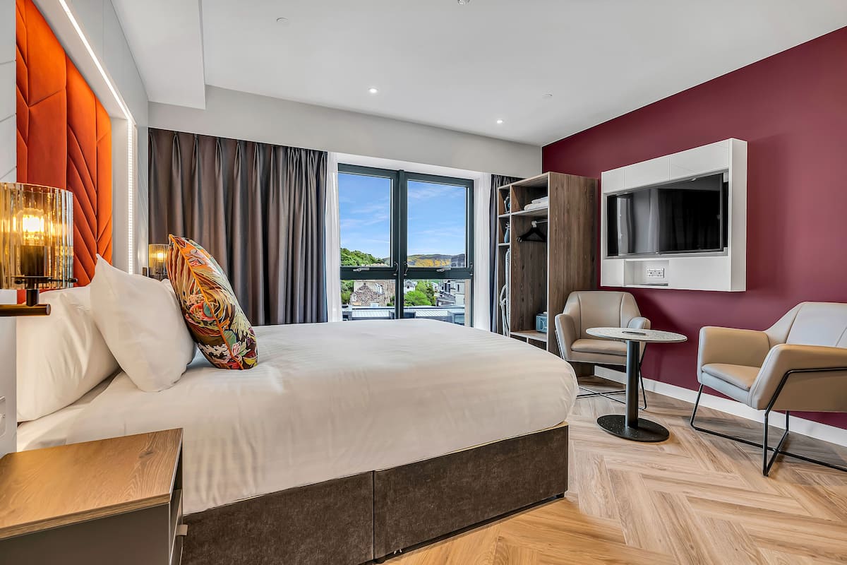 A modern hotel room with a large bed, a TV mounted on the wall, and a view of the cityscape through the window.