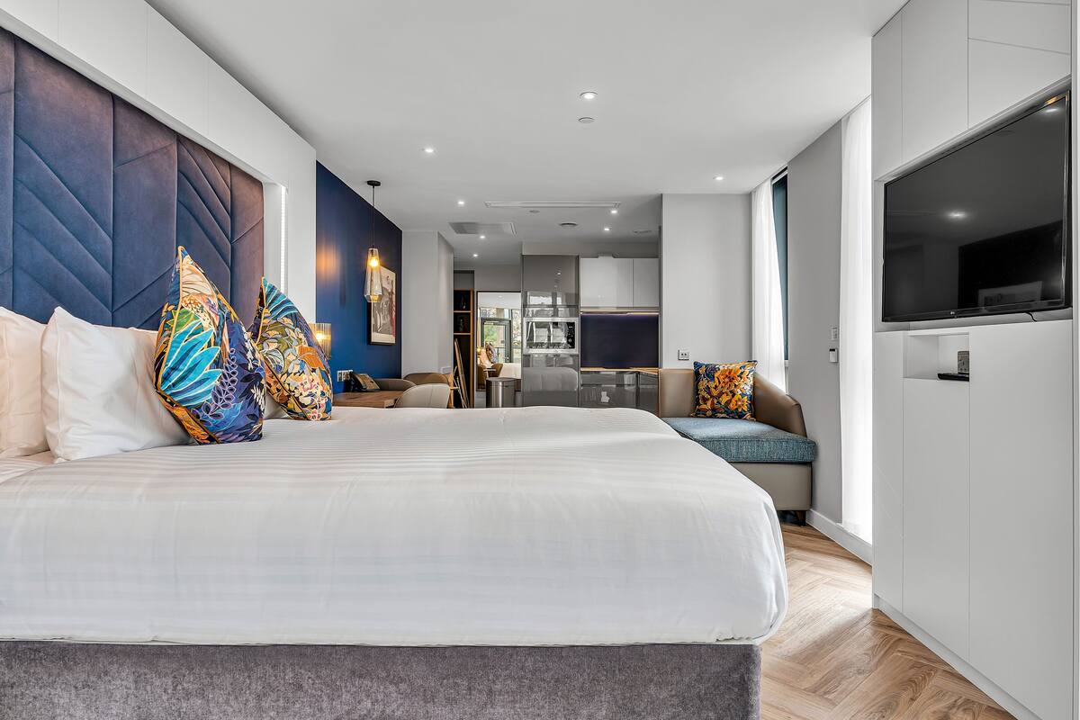 A modern hotel room with a large bed, a sofa, a TV, and a kitchenette.