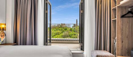 Habitación de hotel con una cama grande, un banco, un armario de madera y un balcón con vistas a un paisaje urbano.