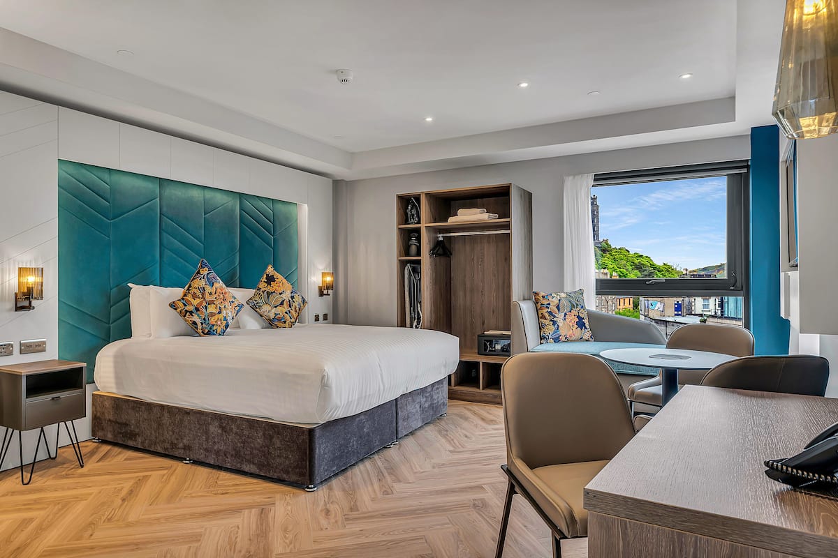 A modern hotel room with a large bed, a seating area with a table, and a wardrobe.