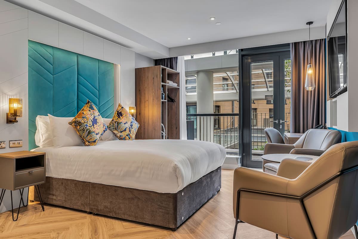 A modern hotel room with a large bed, a wooden nightstand, a seating area with a chair, and a view of the outside through a glass door.