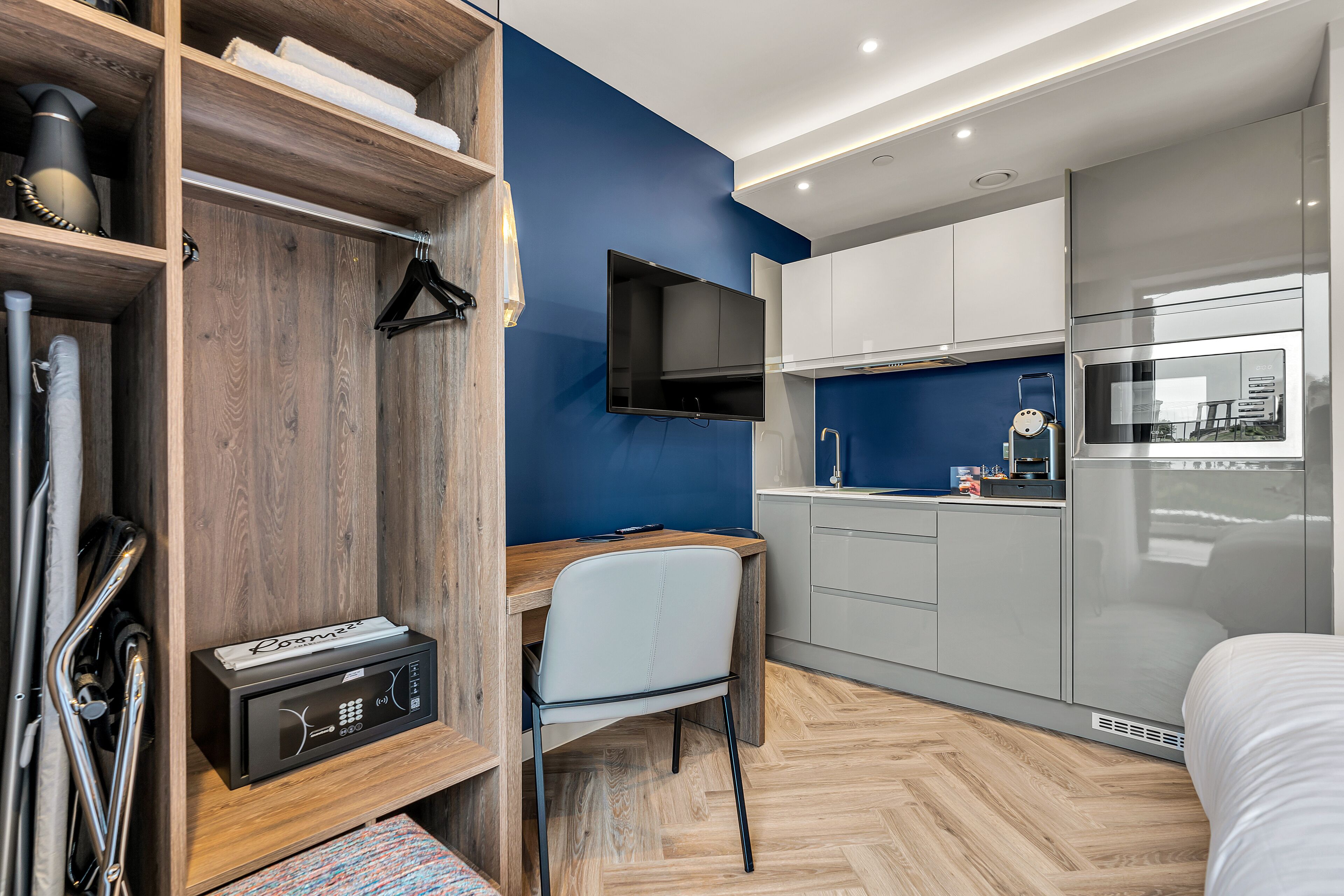 A modern hotel room with a wooden desk, a chair, a flat-screen TV mounted on the wall, a kitchenette with a fridge and microwave, and a bed with white bedding.