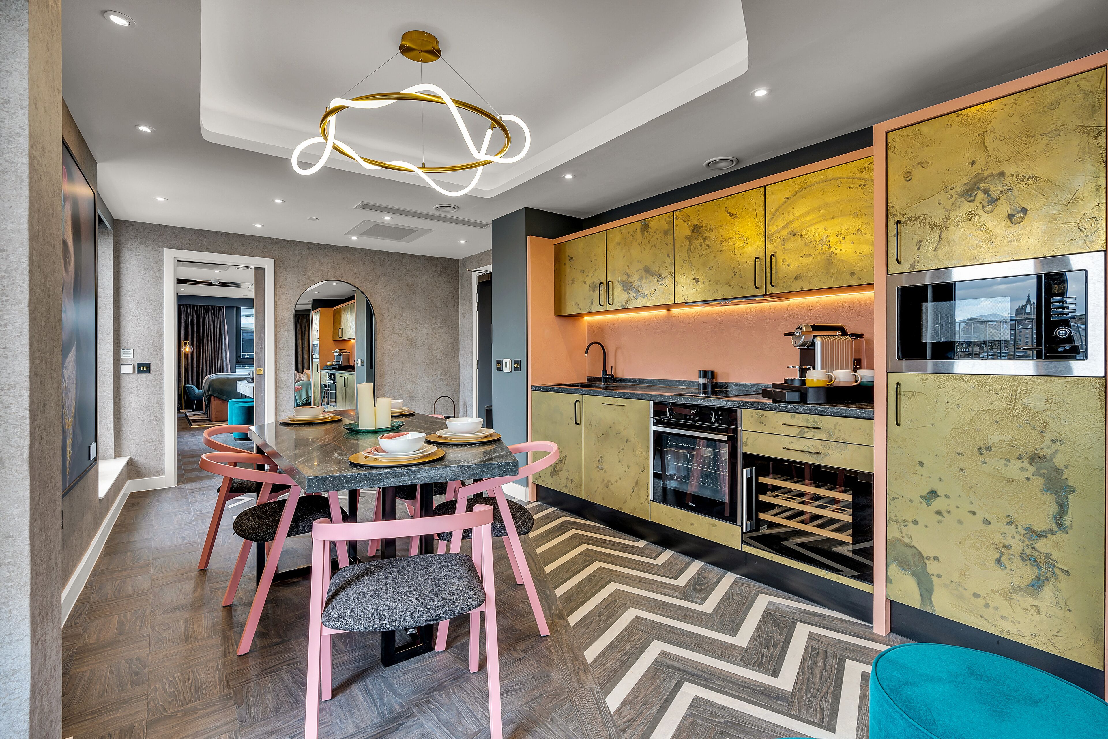 A modern kitchen with a dining area, featuring a herringbone-patterned floor, a central island, and pink chairs.