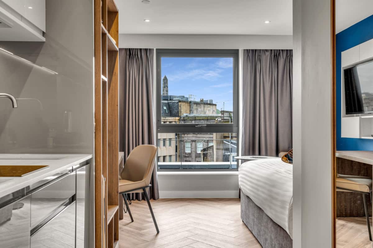 A modern hotel room with a large window, a bed, a desk, and a chair.