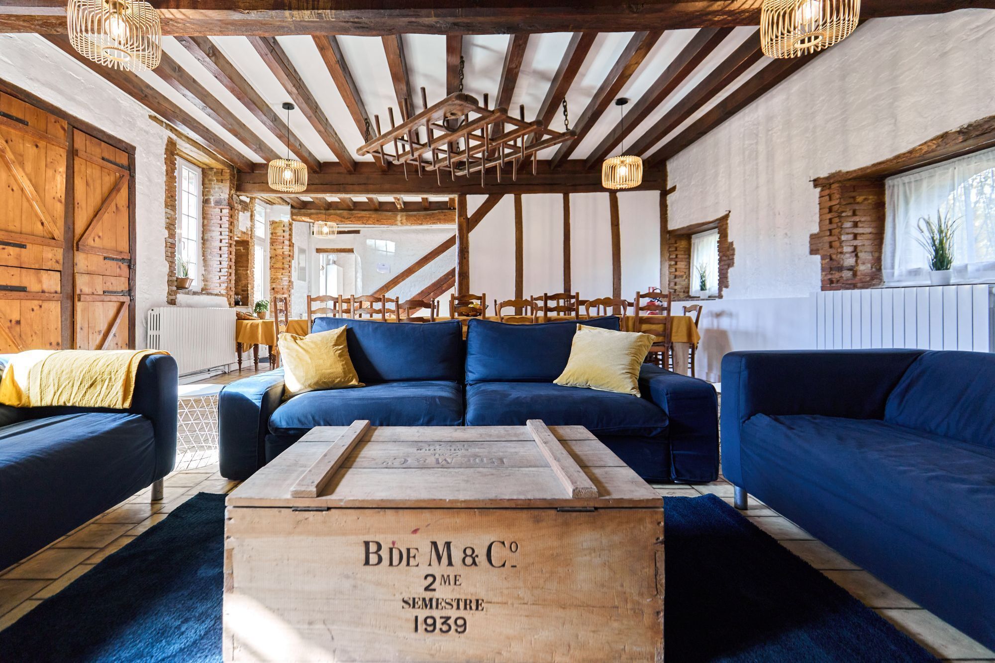 Un intérieur rustique avec des poutres apparentes, un canapé bleu et une vieille malle en bois portant l’inscription « Bde M&C 2e semestre 1939 ».