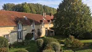 A two-story house with a red-tiled roof, a garden with various shrubs and a large tree, and a clear sky. - Gîte 14 people - 5 bedrooms - 5 bathrooms - Large spaces (Fontaine-sur-Ay)