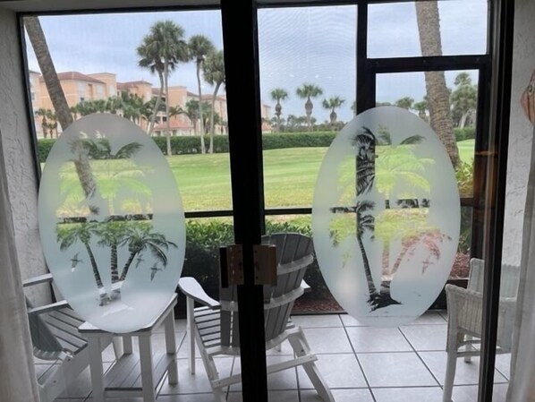 Une pièce avec deux chaises blanches et une table, une fenêtre avec des panneaux de verre dépoli ornés de motifs de palmiers, et une vue sur une pelouse verdoyante et des bâtiments.