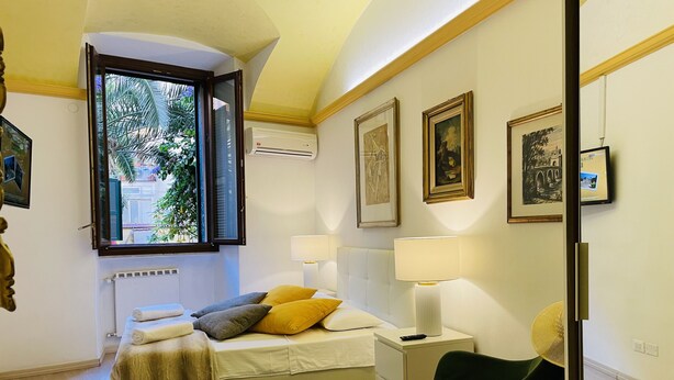 A bedroom with a bed, bedside lamps, a chair, a window with an air conditioner, and framed pictures on the wall.