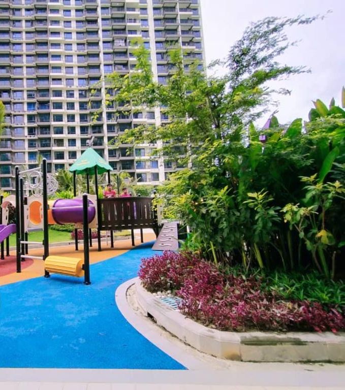 Un parque infantil con toboganes y una estructura para escalar, rodeado de follaje y una zona de juegos azul, con edificios altos al fondo.