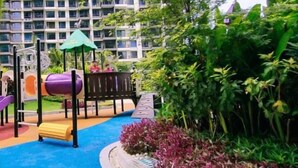 Un parque infantil con toboganes y una estructura para escalar, rodeado de follaje y una zona de juegos azul, con edificios altos al fondo.