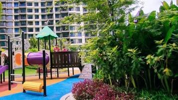 Un parque infantil con toboganes y una estructura para escalar, rodeado de follaje y una zona de juegos azul, con edificios altos al fondo.