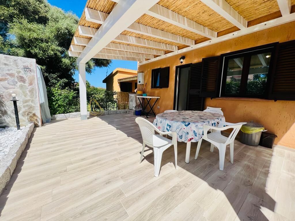 An outdoor patio with a round table and white chairs, a stone wall, and a wooden pergola.