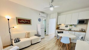 A modern kitchen with white cabinets, a microwave, and a stove. There is a white sofa with a black and white pillow, a small table with chairs, and a ceiling fan. - Residence Oasi La Cinta Centro (San Teodoro)
