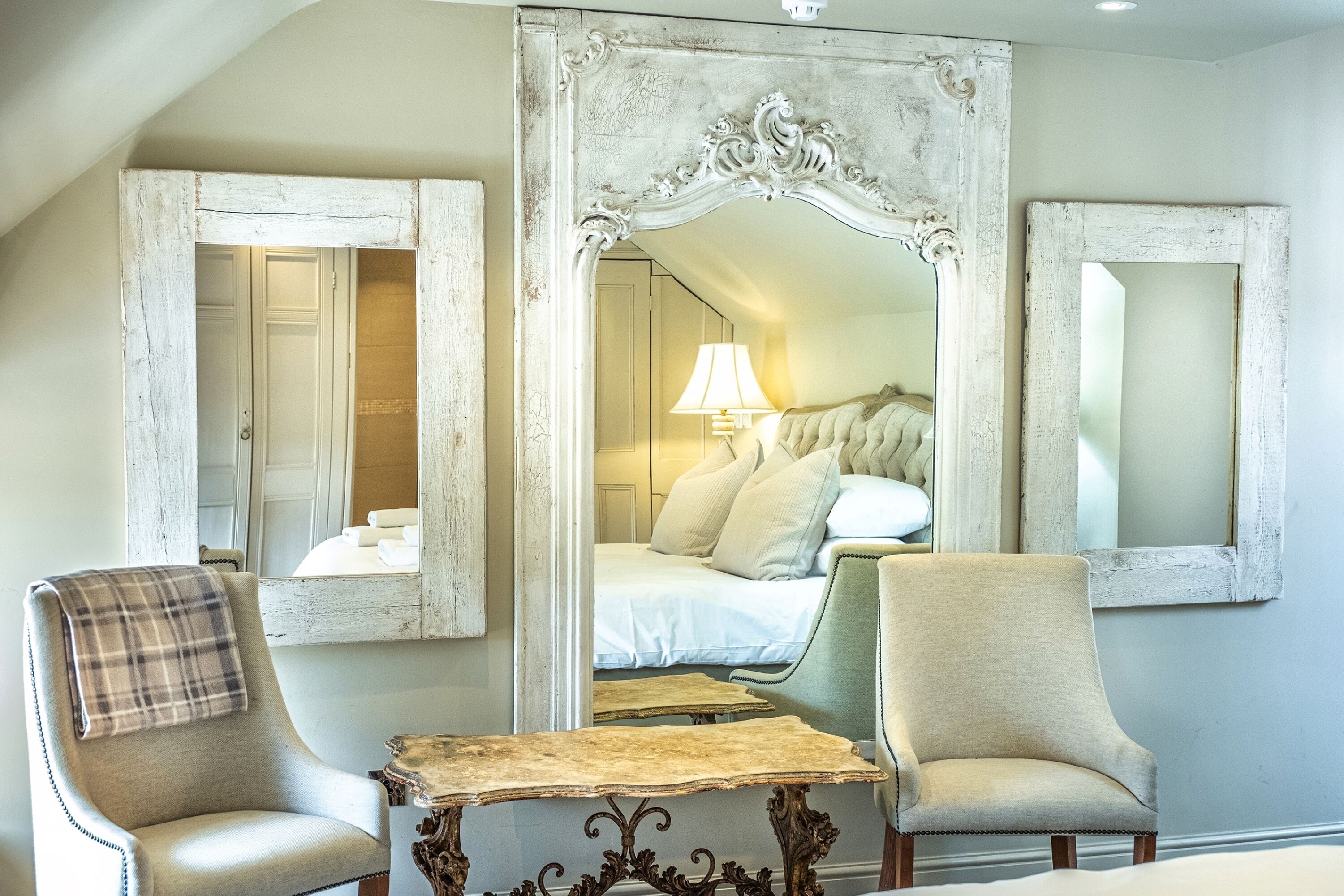 A bedroom with a large mirror, two chairs, a small table, and a bed with a lamp.