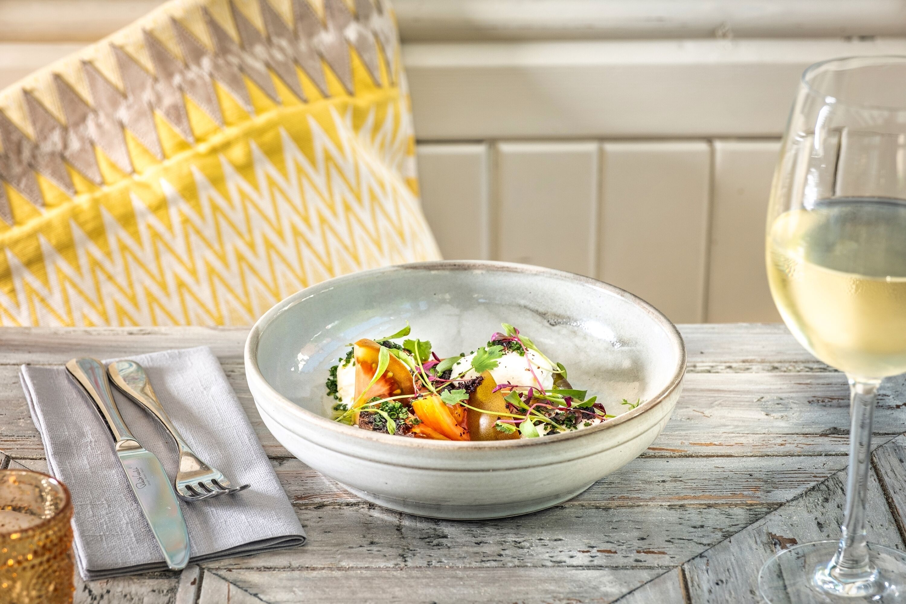 A bowl of food on a rustic wooden table with a glass of white wine and cutlery.