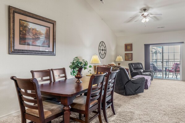 A dining area with a wooden table and chairs, a framed painting, a clock, and a view through a glass door.