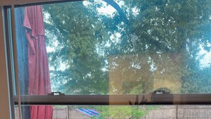 A window with a view of a backyard, a red cloth hanging on the left, and a white air conditioning unit below. - Private, Clean and Charming at amazing rate (Manchester)