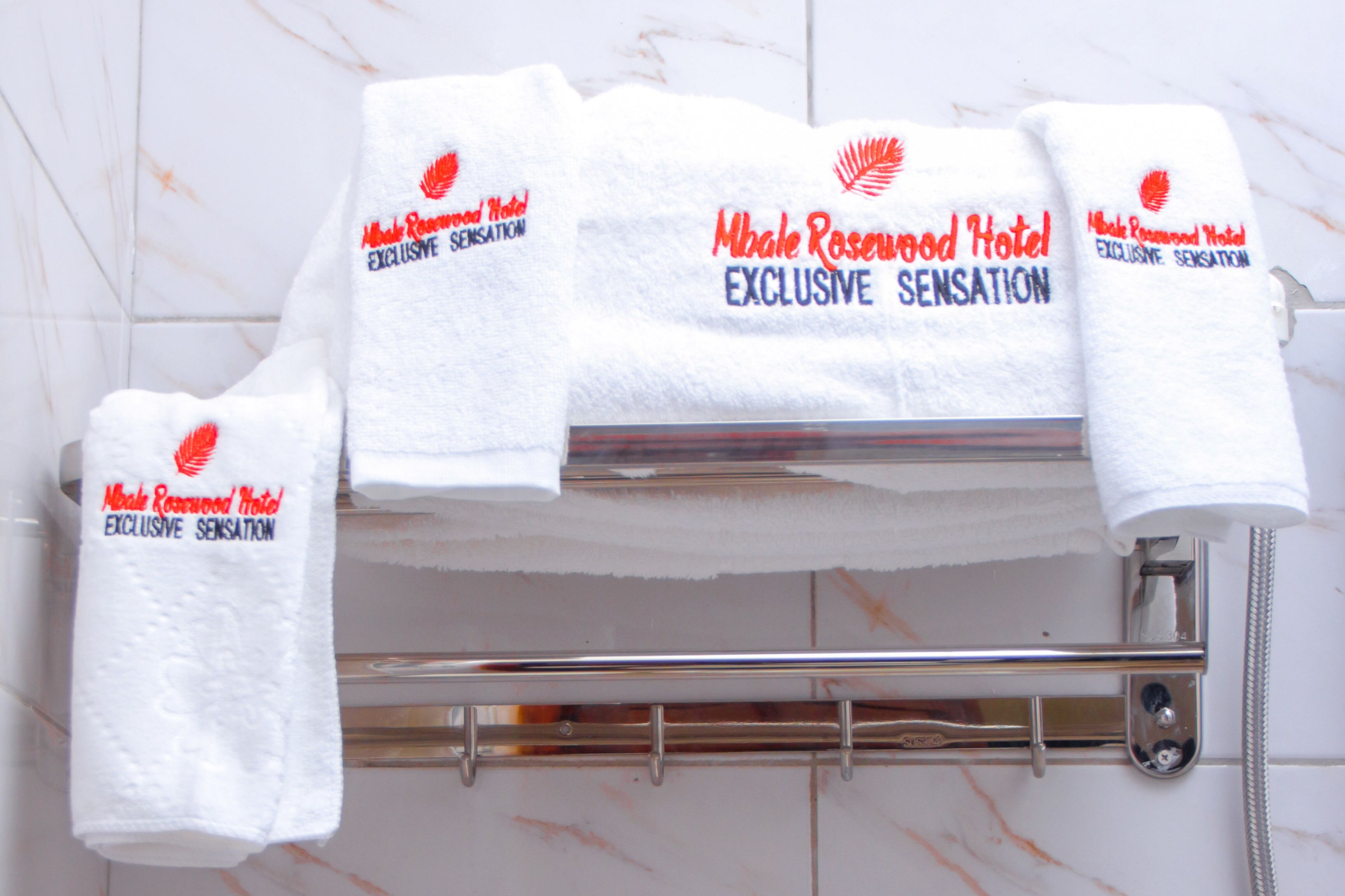 A set of white towels with red text and a logo, hanging on a metal towel rack in a bathroom with white marble tiles.