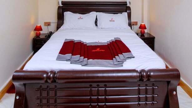 Una cama bien hecha con un dosel blanco, dos lámparas rojas sobre mesitas de noche y un cartel que dice 'Mbale Rosewood Hotel Exclusive Sensation'.