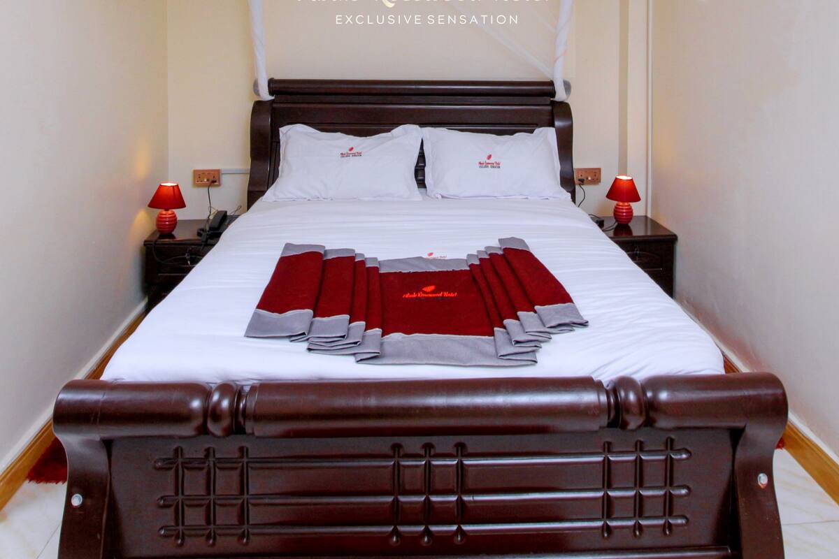 A neatly made bed with a white canopy, two red lamps on nightstands, and a sign reading "Mbale Rosewood Hotel Exclusive Sensation".