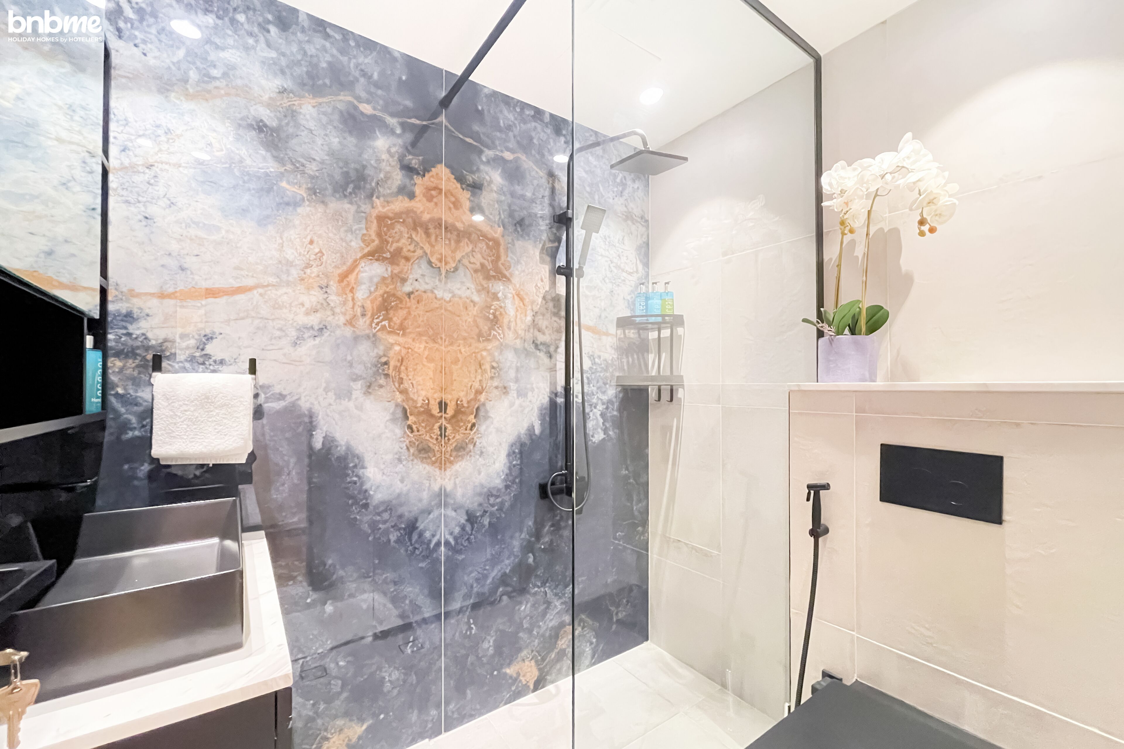 Un baño moderno con ducha de vidrio, paredes de mármol, tocador con lavamanos y una orquídea en maceta.