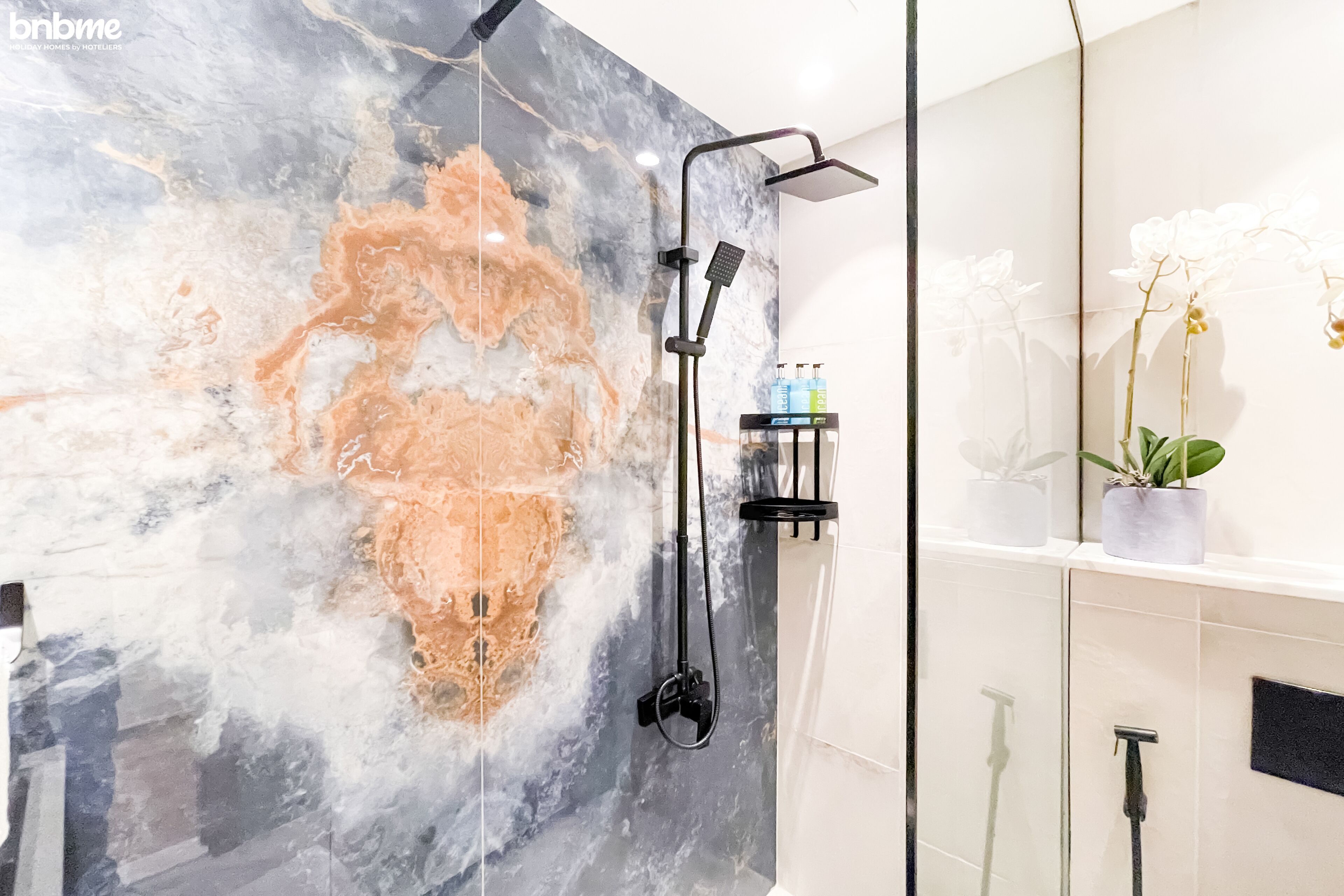 A modern bathroom with a large marble wall, a black showerhead, and a potted orchid on the shelf.