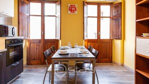Ein Essbereich mit Holztisch, gedeckt für vier Personen, flankiert von großen Fenstern mit Holzläden, eine Küche mit modernen Geräten und eine gelbe Wand mit einer Uhr.