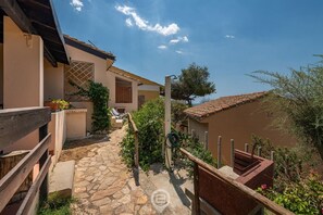 Un chemin de pierres mène à un bâtiment doté d’un toit en tuiles, entouré de verdure et délimité par une clôture en bois.