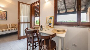 A dining area with a wooden table, two chairs, a small white table, and a window with a view of greenery. - Porto luna 24 - Selene (Villasimius)