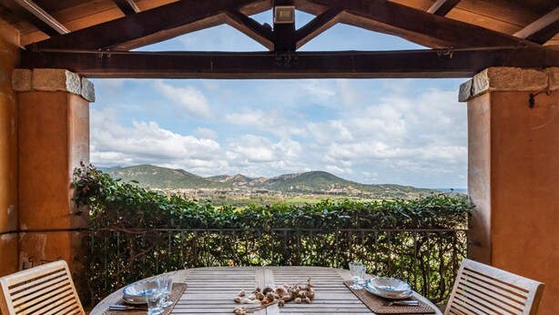 A wooden table set for two with plates, glasses, and a centerpiece of garlic bulbs, overlooking a mountainous landscape. - Casa Perla - Domus de Maria (Domus De Maria)