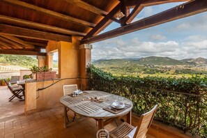 An outdoor patio with a wooden table and chairs, a stone bench, and a view of a green landscape with hills. - Casa Perla - Domus de Maria (Domus De Maria)