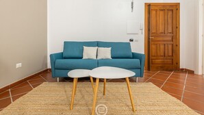Quarto com sofá azul, duas mesinhas brancas, tapete tecido e porta de madeira.