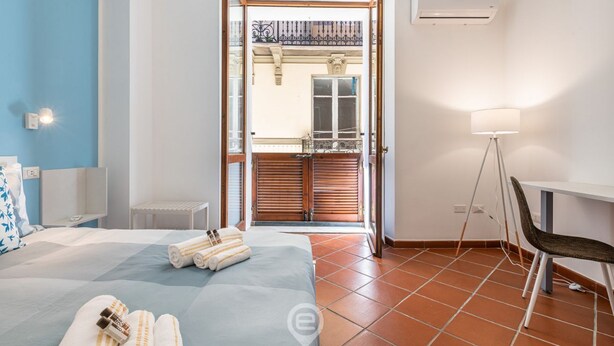 Quarto de hotel com cama, escrivaninha, cadeira, luminária e vista para a varanda externa.