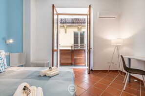 A hotel room with a bed, a desk, a chair, a lamp, and a view of the outside balcony. - Elisabetta Apartment (Cagliari)