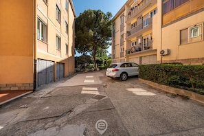 A residential street with apartment buildings, a parked car, and a hedge.