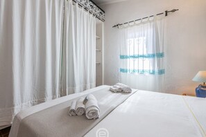 A hotel room with a bed, white curtains, a window with blue accents, and a lamp.