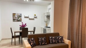 A modern living room with a brown sofa, a dining table with chairs, and a kitchen area with white cabinets and a stove.