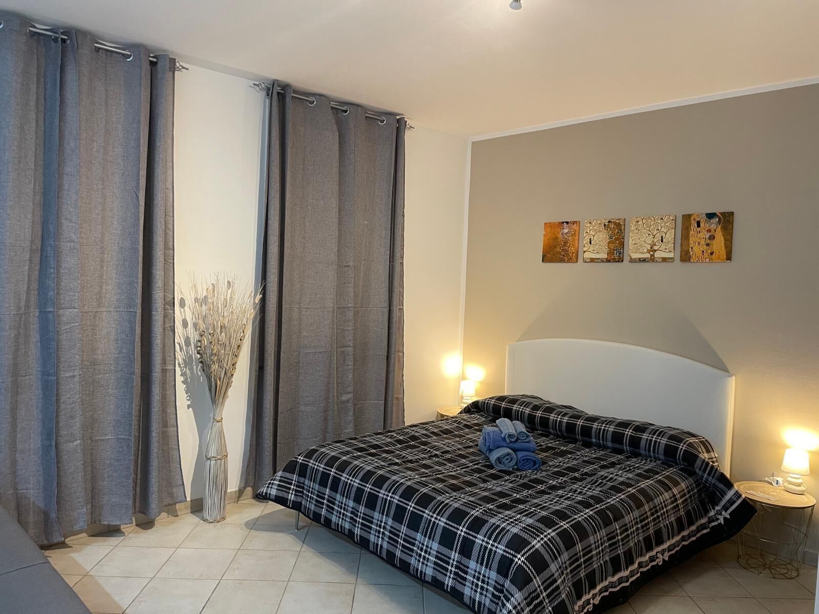 A bedroom with a bed, curtains, a vase with dried plants, and wall art.