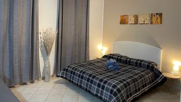 Un dormitorio con cama, cortinas, un jarrón con plantas secas y decoración en la pared.