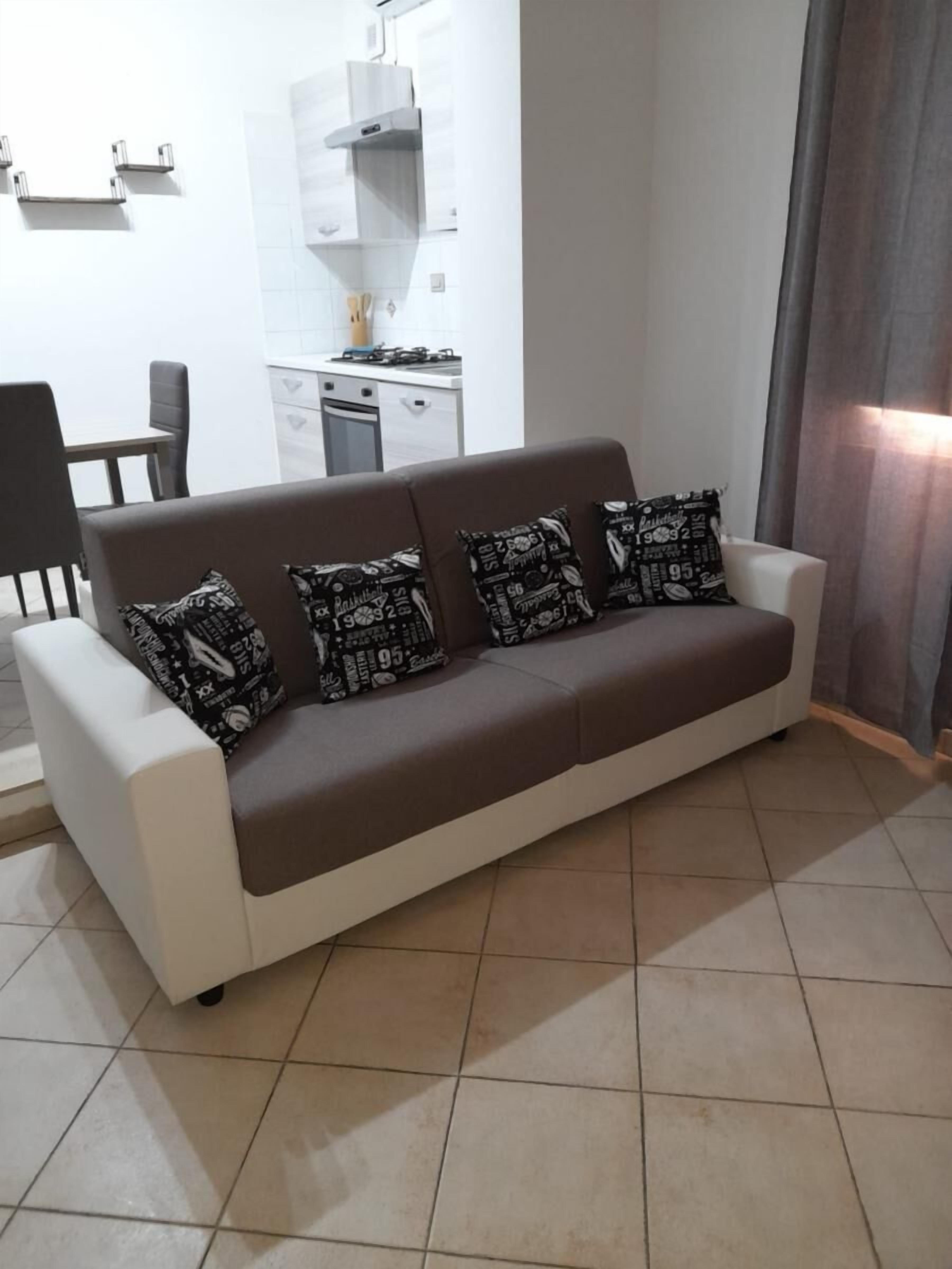 A modern living room with a white and brown sofa, four patterned cushions, a dining area with a table and chairs, and a kitchen in the background.